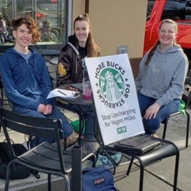 Three students sitting outside starbucks with sign that says stop upcharging for vegan milks