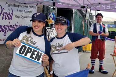 Two women in baseball shirts and caps, one of them holding an open sign, and a clown standing in the background