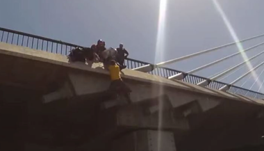 people holding a man who is hanging off of a bridge