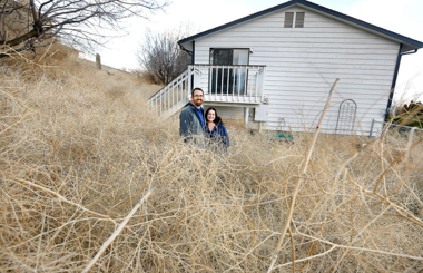 A man and a woman stanfing in front of a house surrounded by tumbleweeds stacked so high you can only see them from the waist up