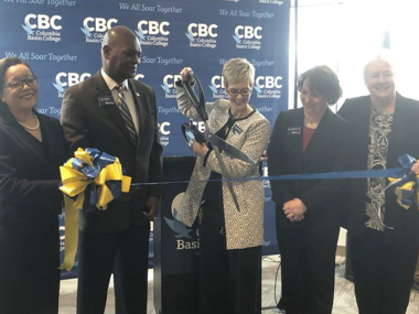 five people standing in a line behind a blue ribbon as the center woman cuts it with giant scissors