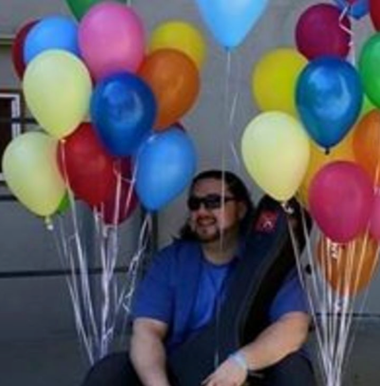 Man (mango) sitting on a porch with a guitar case, shrouded by balloons