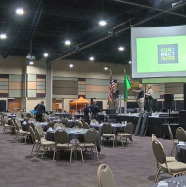 people stand in an an event center with empty tables and the words connect tri cities are projected onto a screen