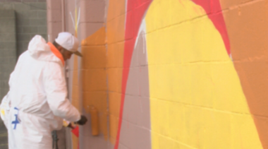 Man in protective, white suit paints large wall of business with roller and orange paint.