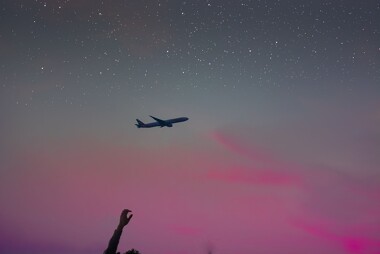 A plane flys overhead while a person reaches a hand up toward the sky