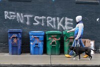 Person walking their dog down a city street with the words rent strike written in grafiiti along the wall of the building next to them